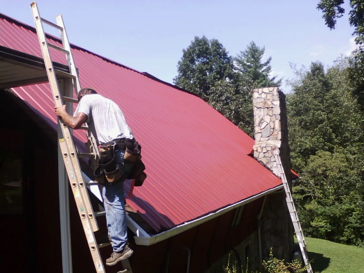 Roofer on a ladder beside a red metal roof and stone chimney — Soffit & Fascia Repair by Roofing Brothers in Tower City